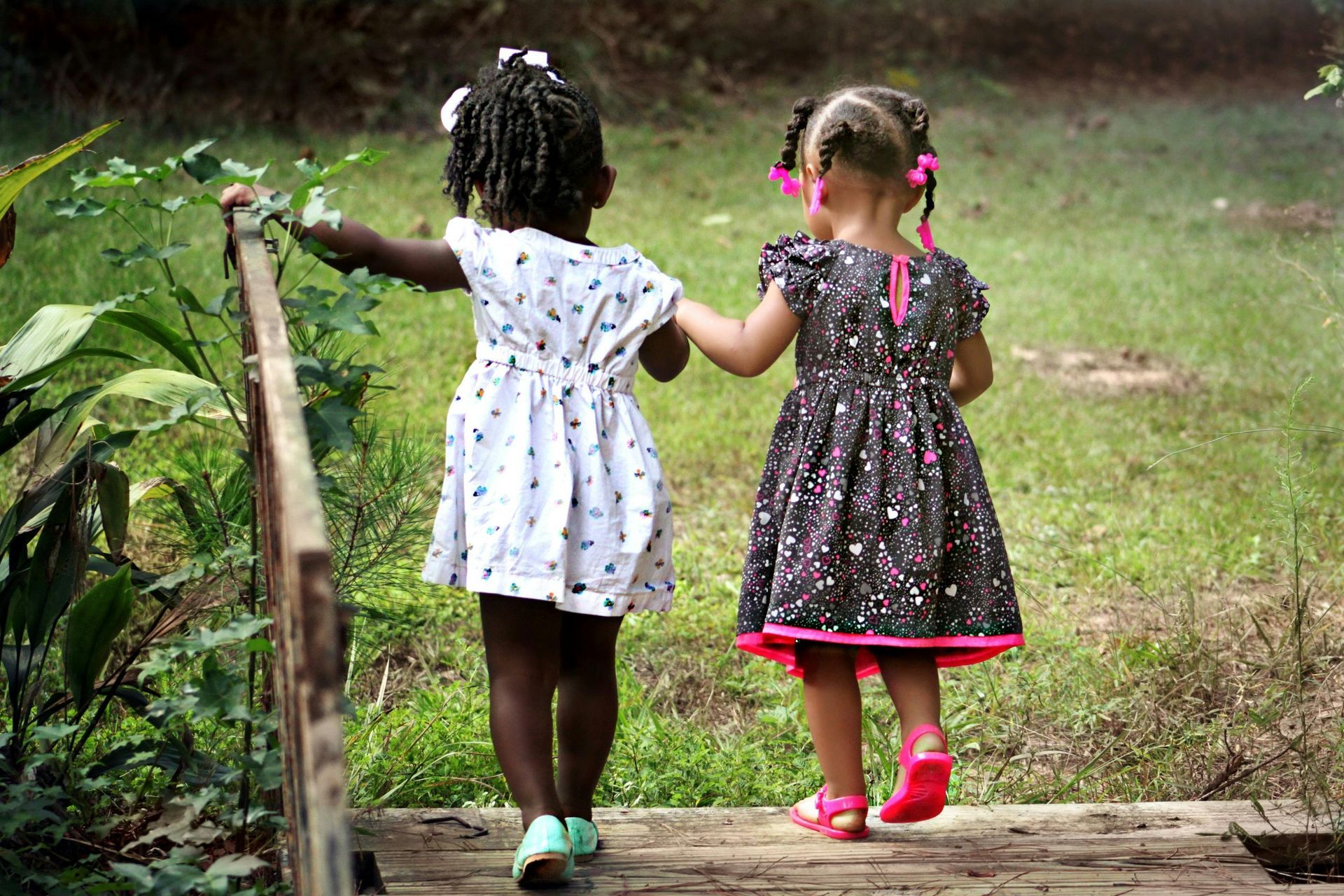 Two young girls in dresses walk hand-in-hand across a wooden bridge outdoors.