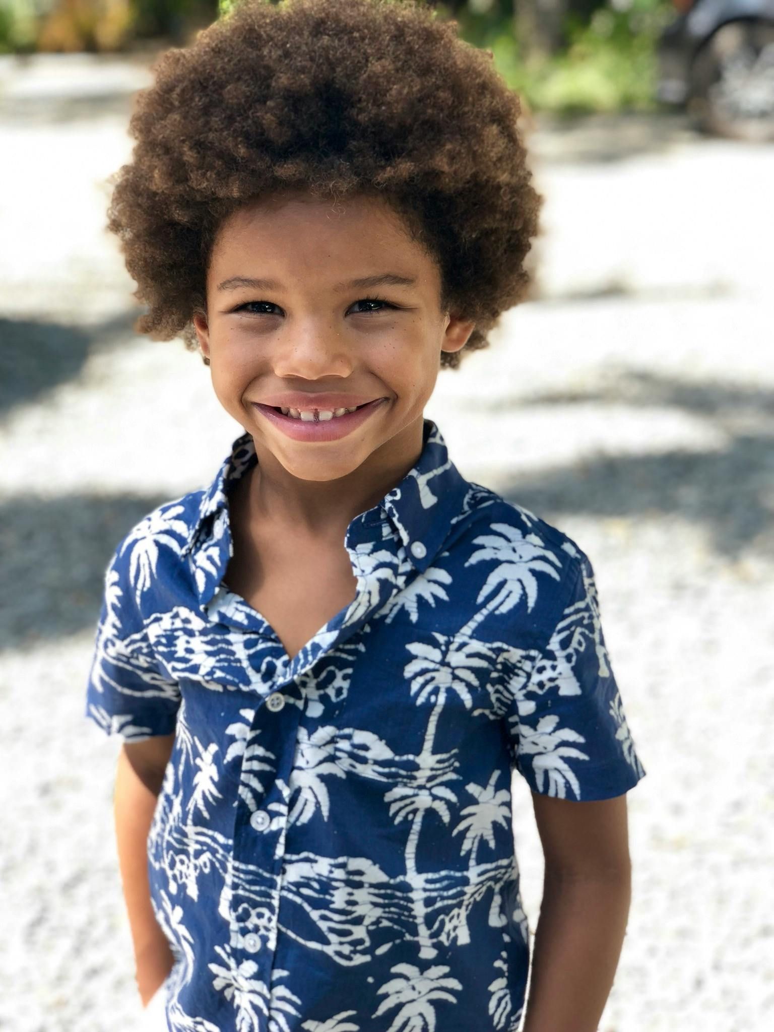 Boy with curly brown hair smiles, wearing a blue palm tree print shirt. Outdoors.