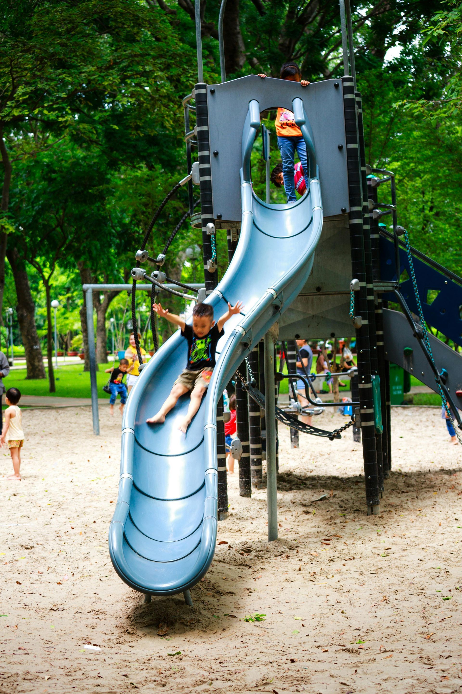Boy slides down a blue slide at a park, smiling with arms outstretched. Other children and trees in the background.