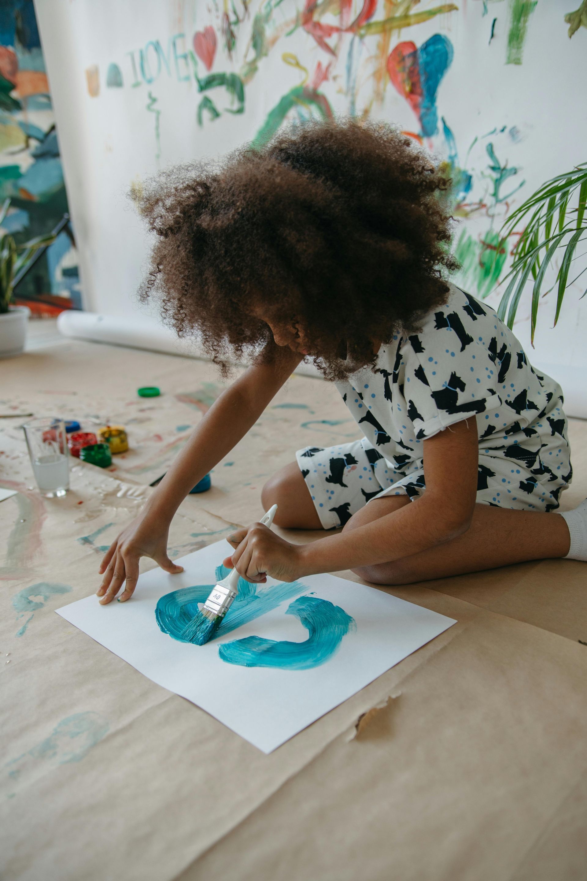 Child kneeling on floor, painting with blue brushstrokes on paper; colorful artwork in background.