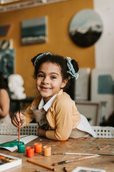 Young child smiles, painting with brush at a table with paints in a studio.