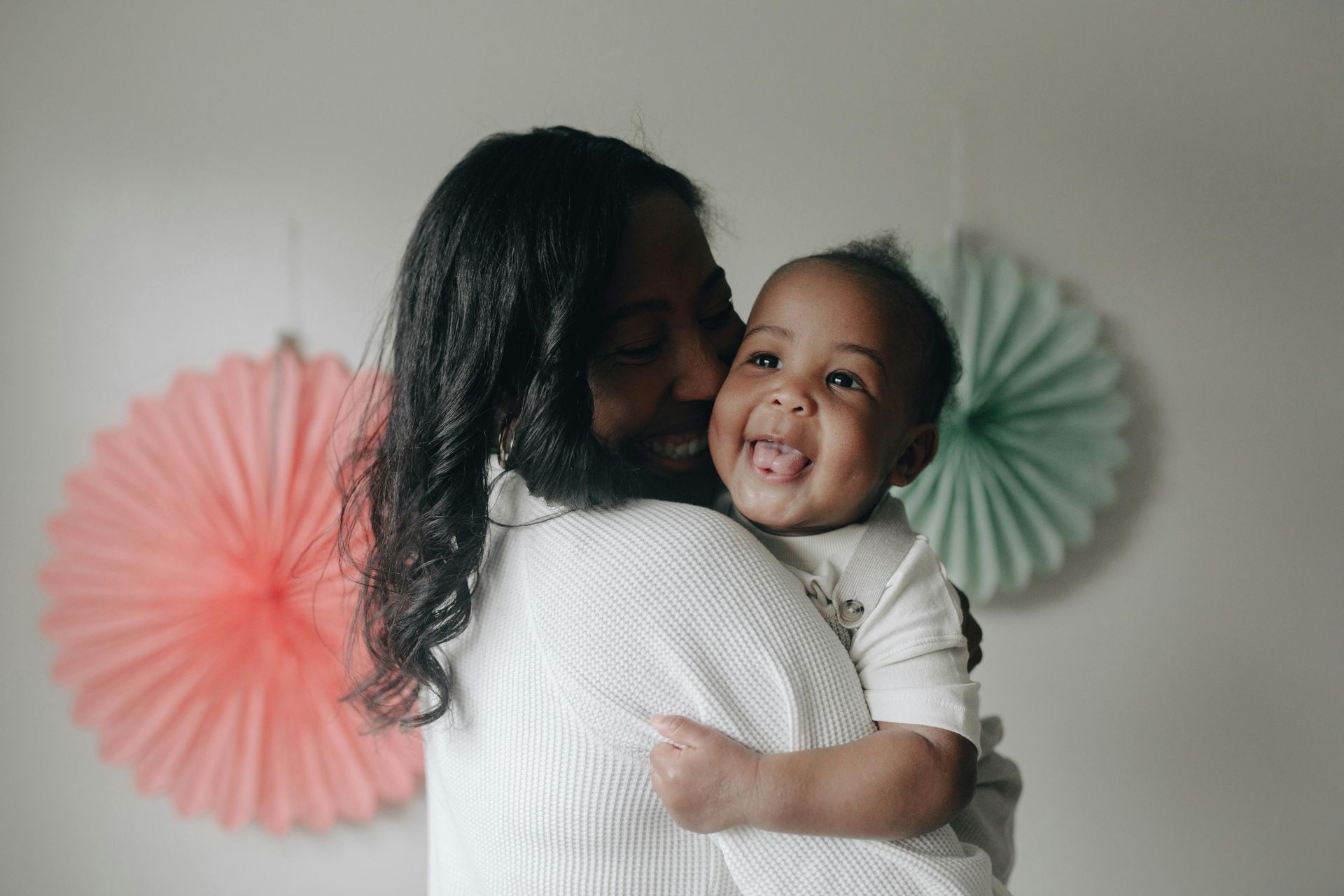 Woman in white sweater holding a smiling baby, kissing cheek. Pastel paper fan decorations in the background.