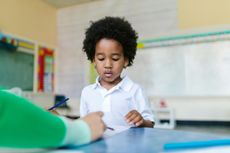 Child looking at paper while someone’s hand points to it in a classroom.