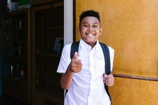 Boy with backpack gives a thumbs-up, smiling near a doorway.