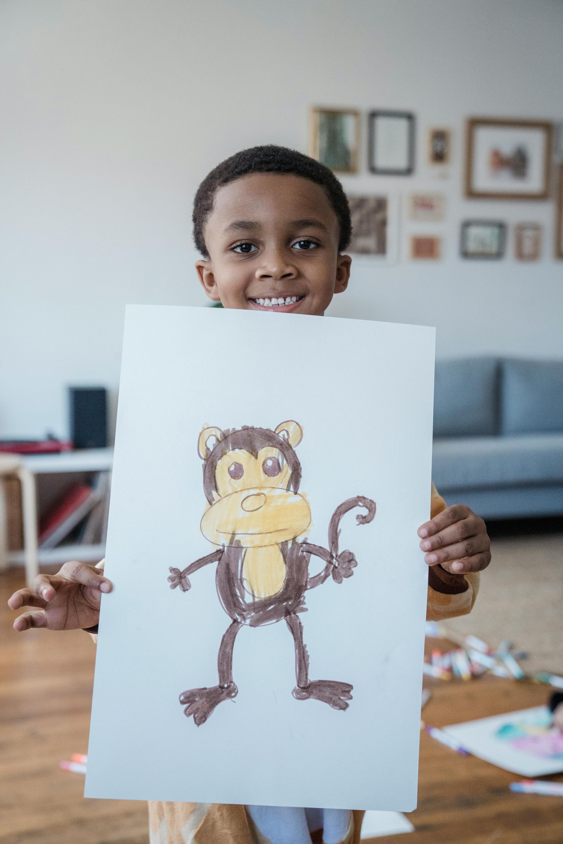 Boy smiling, holding drawing of a monkey. Brown and yellow colors. Indoor setting with art supplies visible.