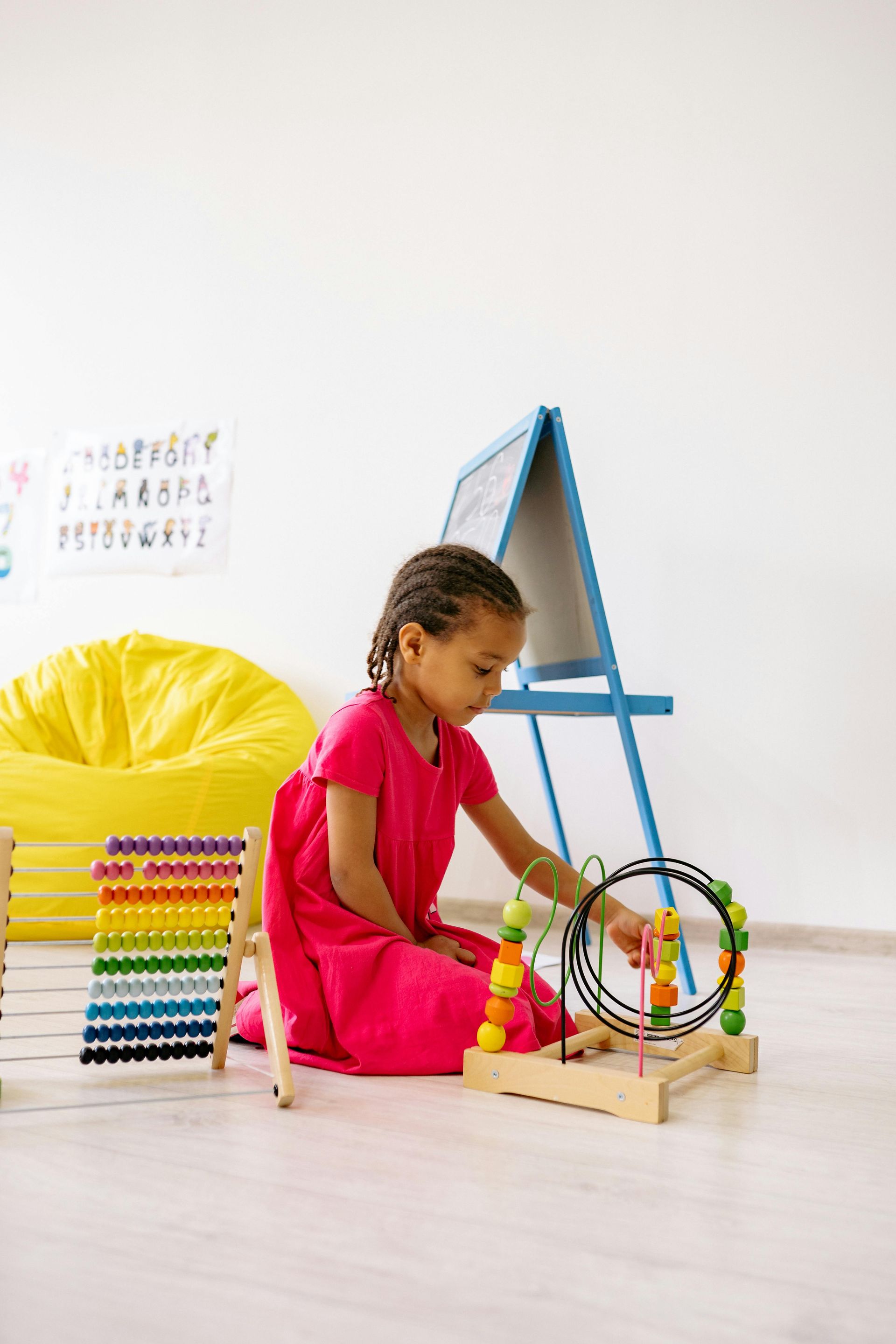 Girl in pink dress playing with a wooden bead maze and abacus on the floor near an easel.