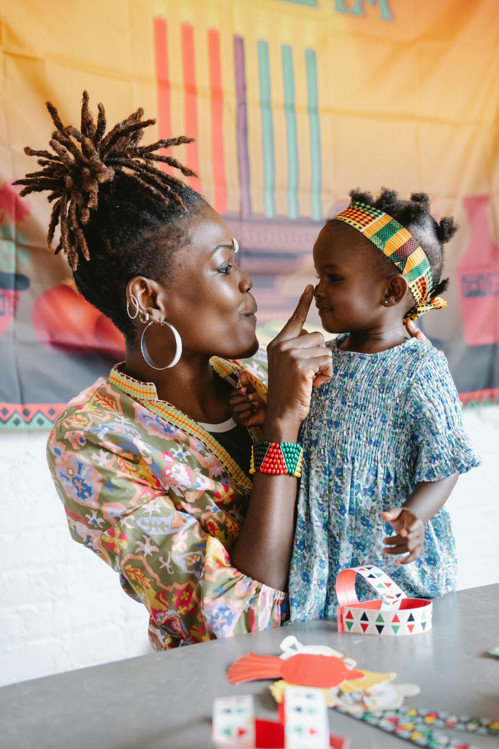 Woman holding child, touching noses. Colorful clothing, craft supplies visible.