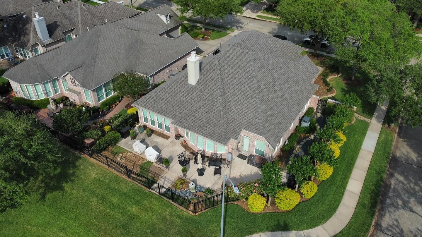 Aerial view of a suburban house with a gray roof, brick exterior, patio, and surrounding landscaping.