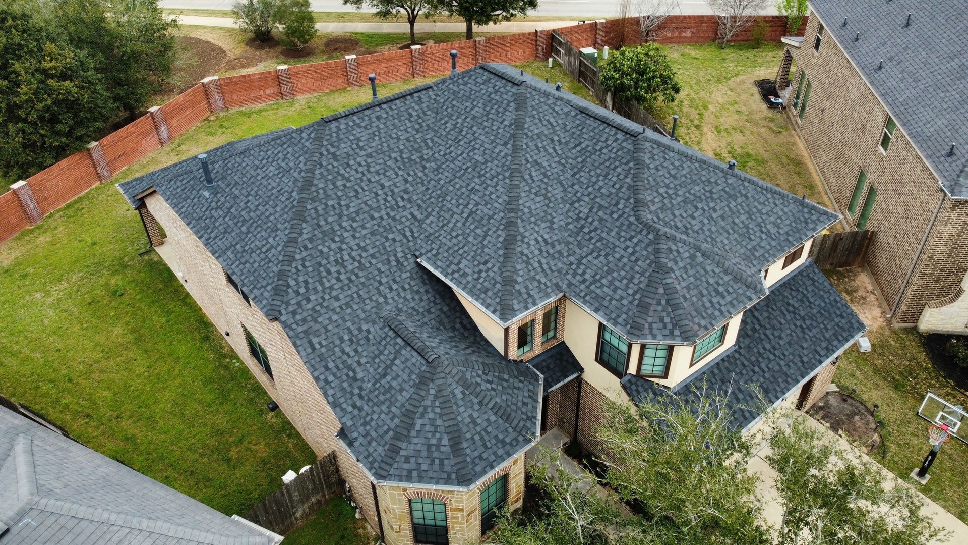 Aerial view of a two-story house with a dark gray shingle roof, green lawn, and a brick fence.