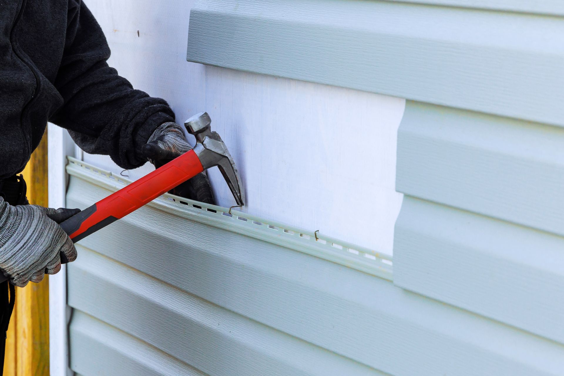 Person wearing gloves using a hammer to remove siding from a wall.