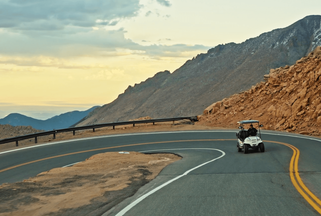 Golf cart on a winding mountain road with a rocky hillside and distant mountains.