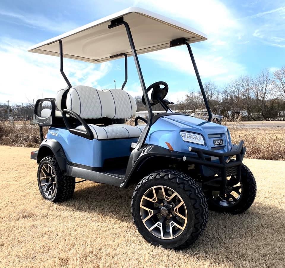 Blue golf cart with white roof, black accents, and off-road tires, parked outdoors on grass.