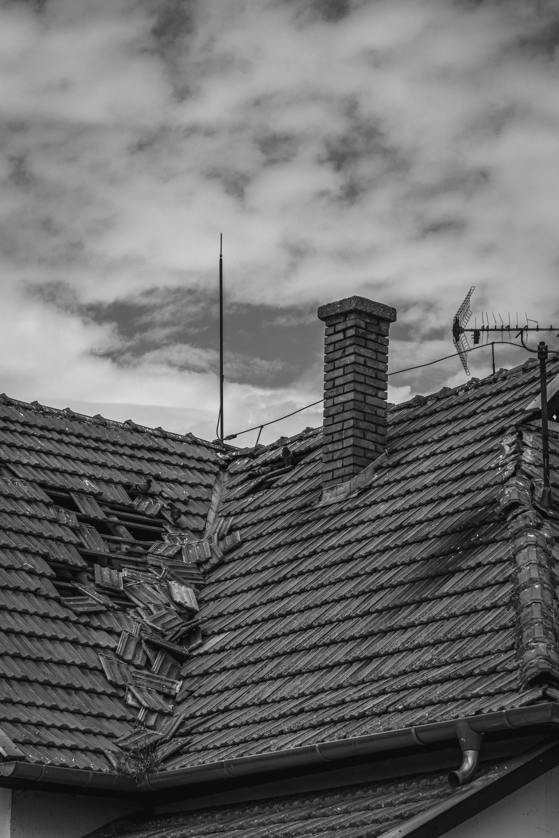 Black-and-white view of a residential tiled roof with significant damage, missing tiles, and a prominent brick chimney.