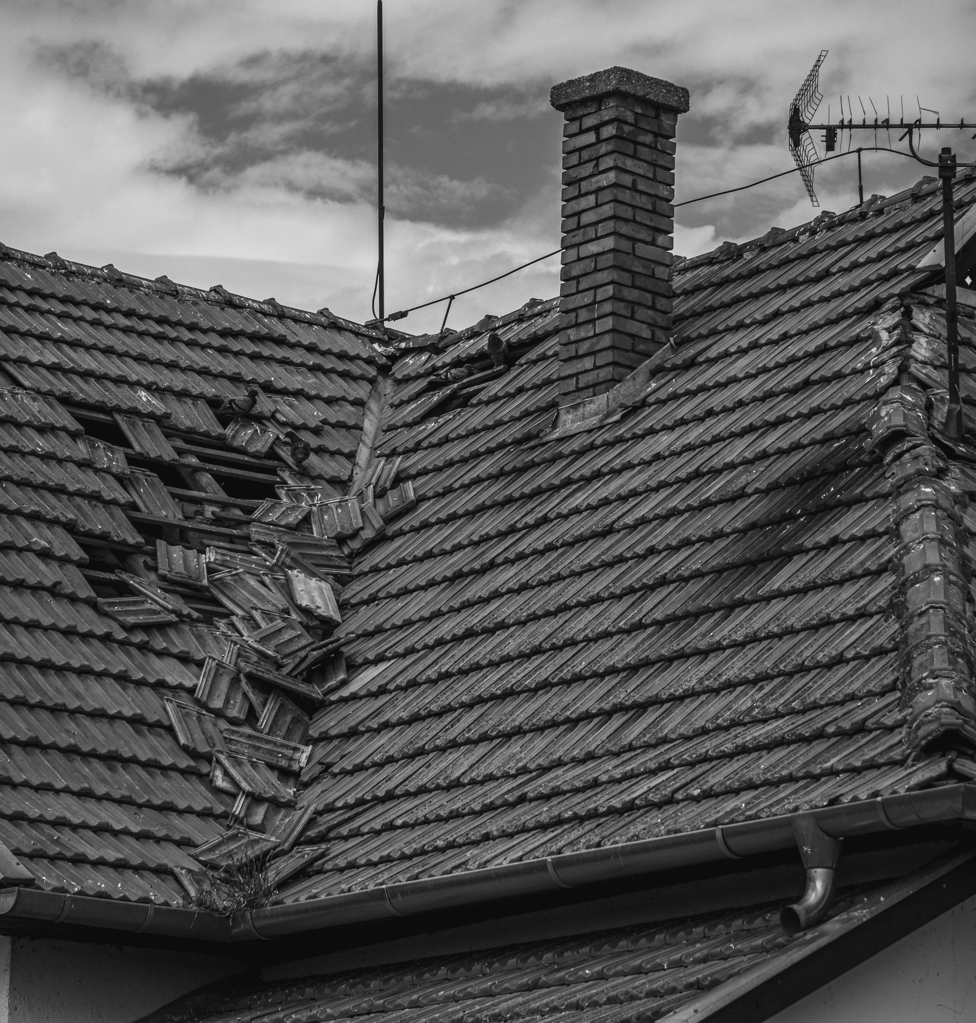 A black and white image of a residential roof with a large section of missing and damaged tiles near a brick chimney.
