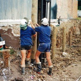 Two workers are driving timber piles into the ground to reinforce the foundation and ensure structural stability. Two workers are driving timber piles into the ground to reinforce the foundation and ensure structural stability.