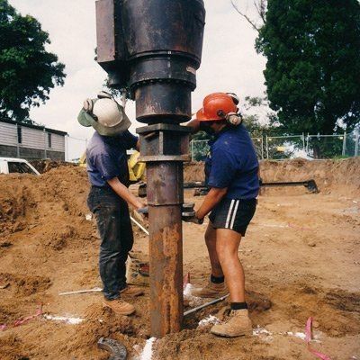 Two construction workers wearing safety gear are performing screw piling at a construction site. Two construction workers wearing safety gear are performing screw piling at a construction site.