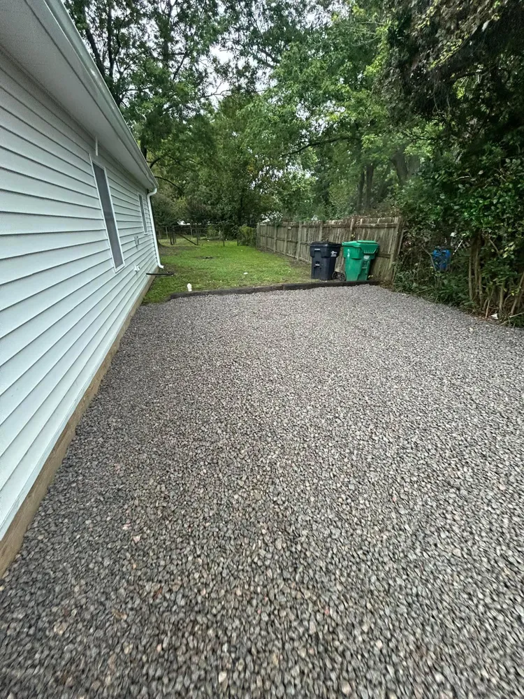 A gravel driveway leading to a house with trash cans in the backyard.