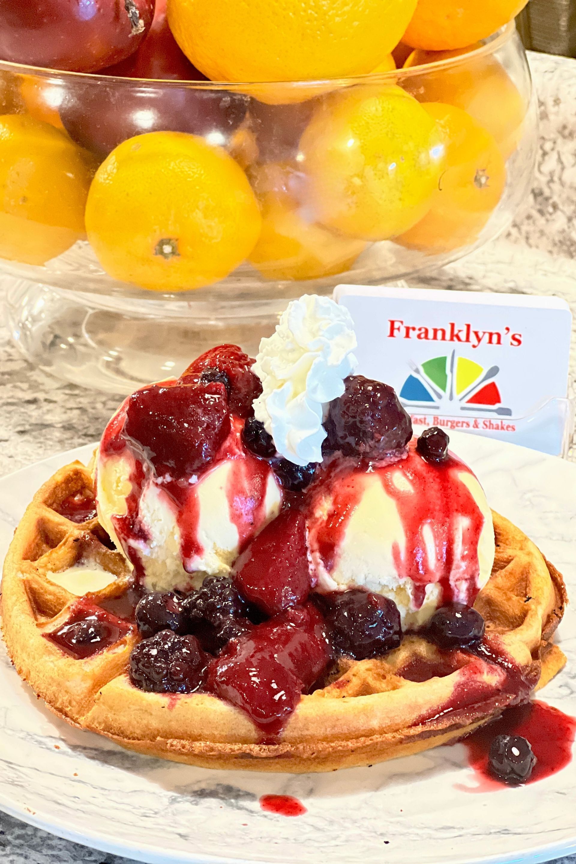 A waffle with ice cream and berries on a plate with a bowl of fruit in the background.