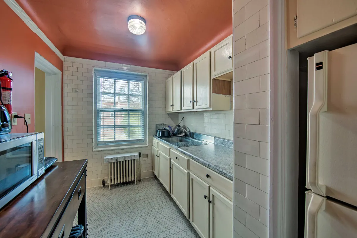 A kitchen with white cabinets , a refrigerator , a microwave and a window.