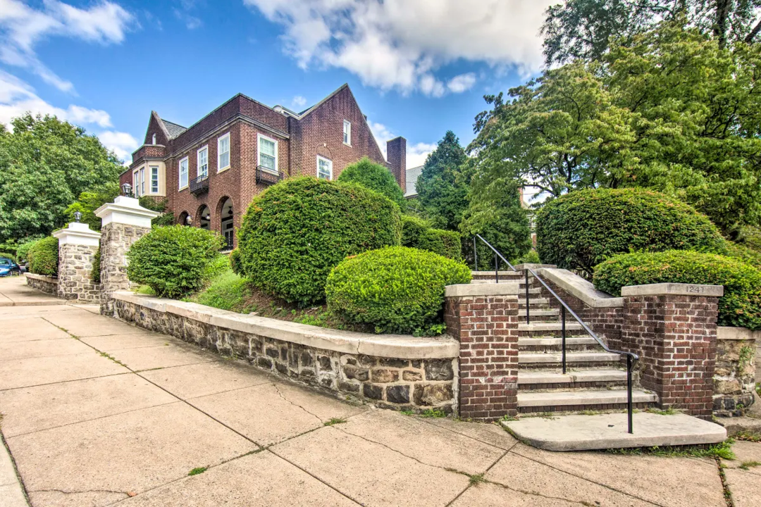 A large brick building with stairs leading up to it is surrounded by bushes and trees.