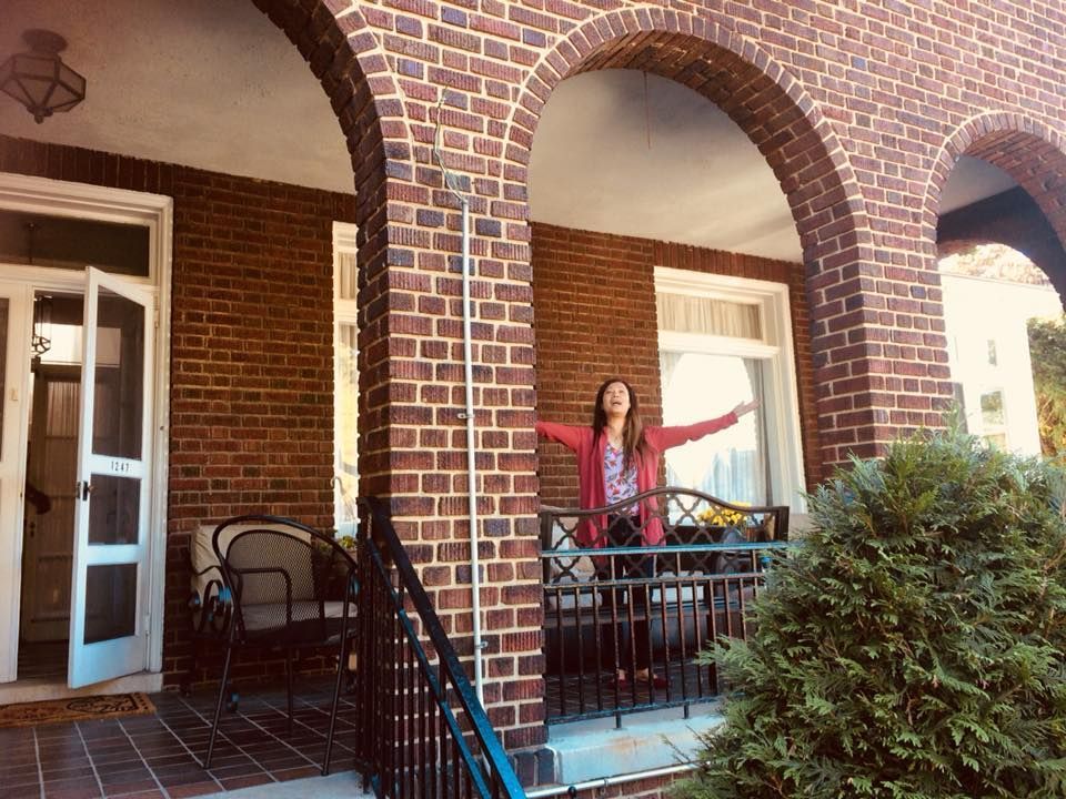 A woman is standing on a porch of a brick house with her arms outstretched.