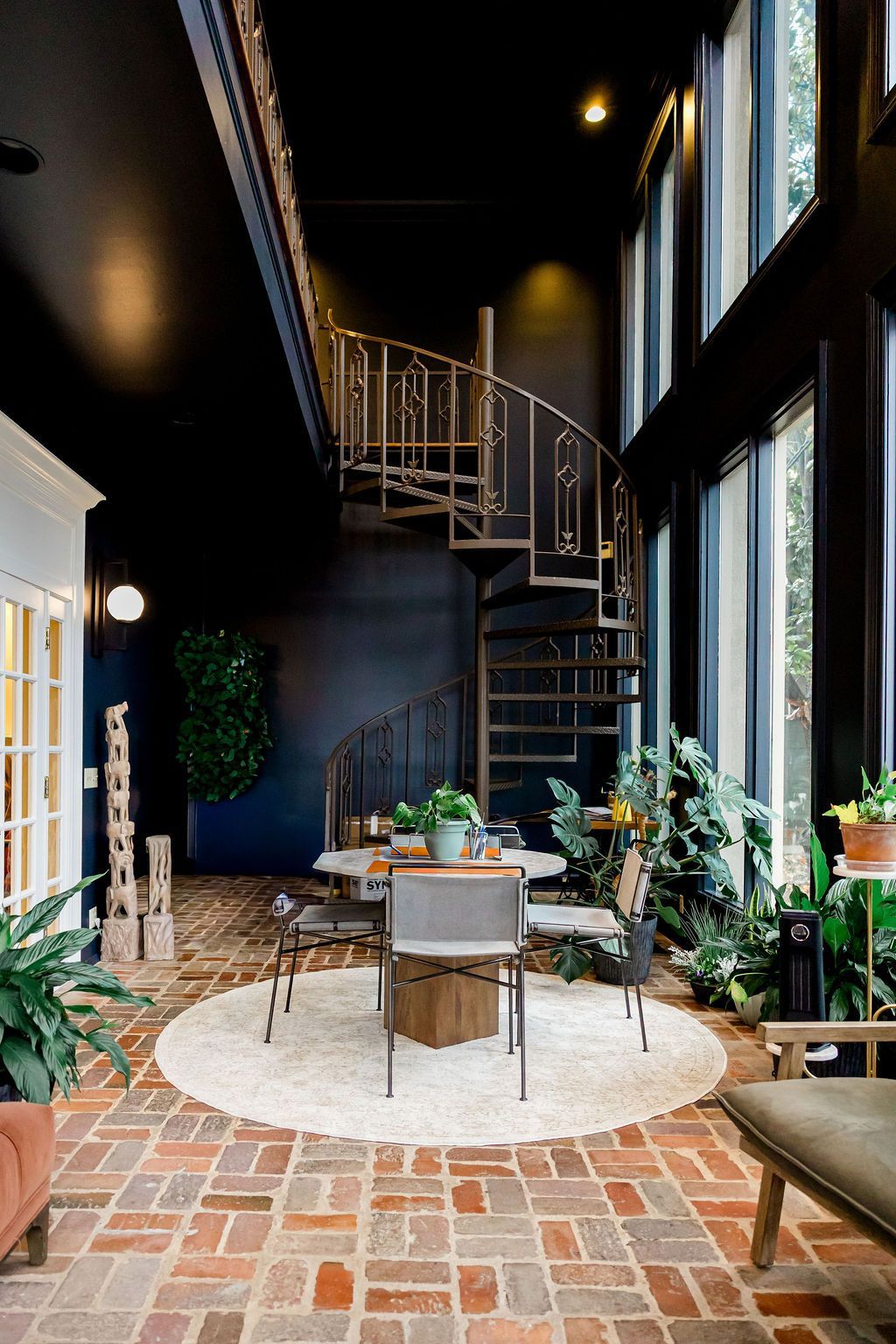 A dining area with a spiral staircase, surrounded by windows and plants. Black ceiling and brick floor. About Us in Nashville, Tennessee