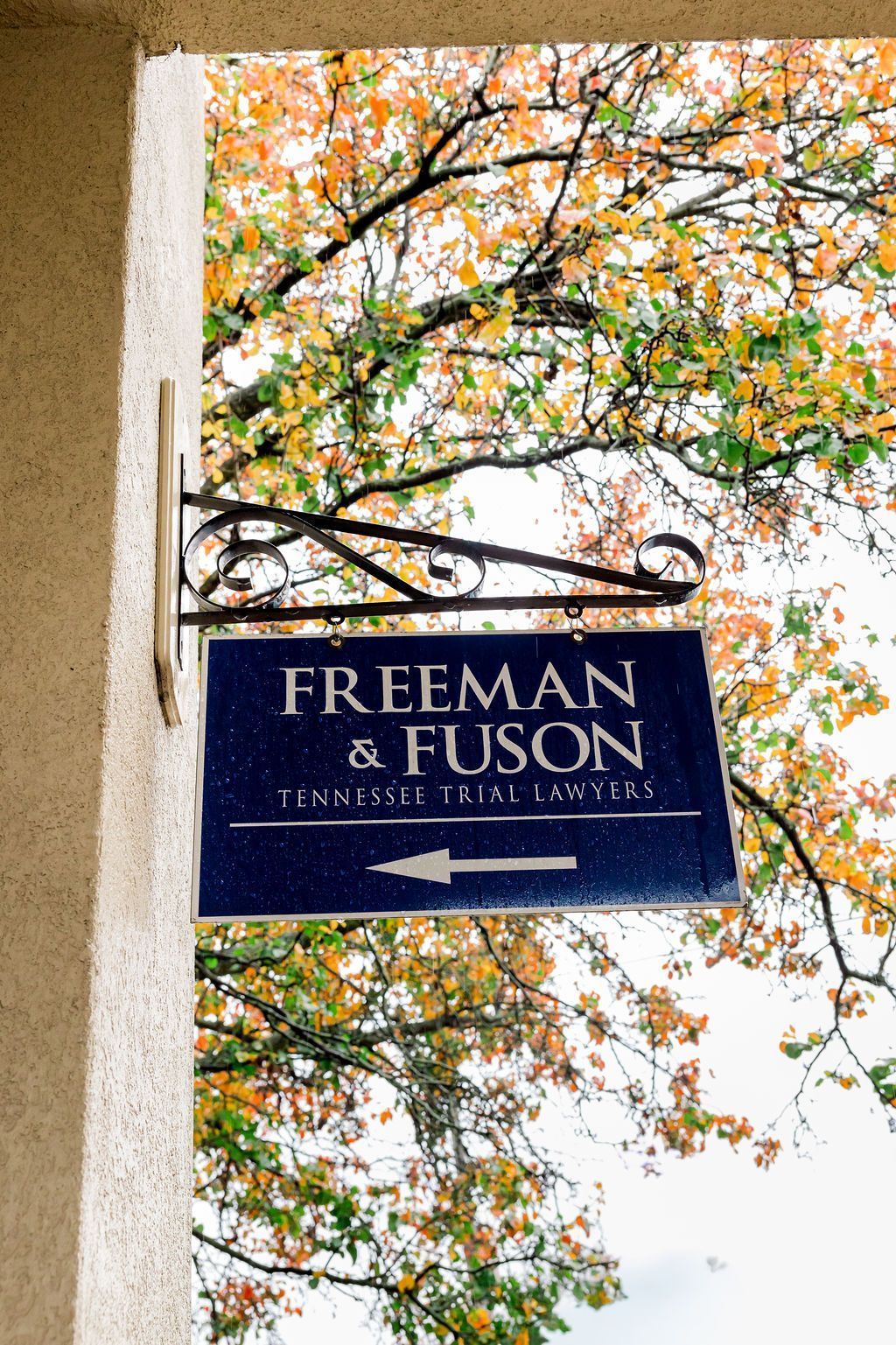 Sign for Freeman & Fuson, a legal firm. Blue sign with white lettering and an arrow. Over a doorway, beneath autumn foliage. Freeman & Fuson Trial Lawyers in Nashville, Tennessee