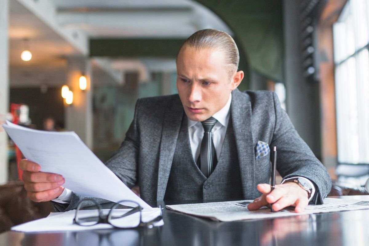 Man in a suit intently reviews documents at a table. He holds a pen and papers in a cafe setting. Forgery in Nashville, Tennessee