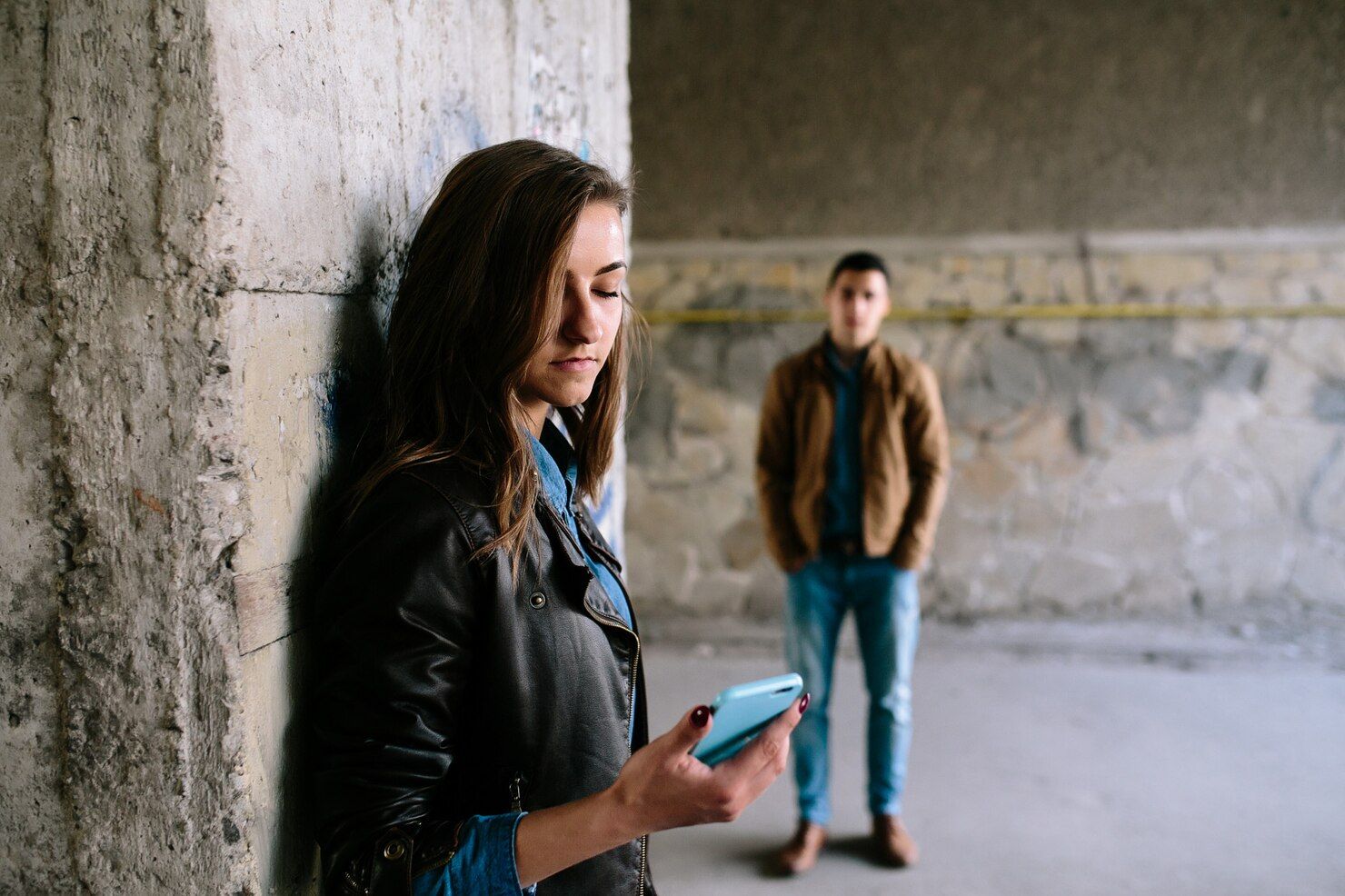 Woman using phone leaning against a concrete wall, man stands in the background.  Stalking in Nashville, Tennessee