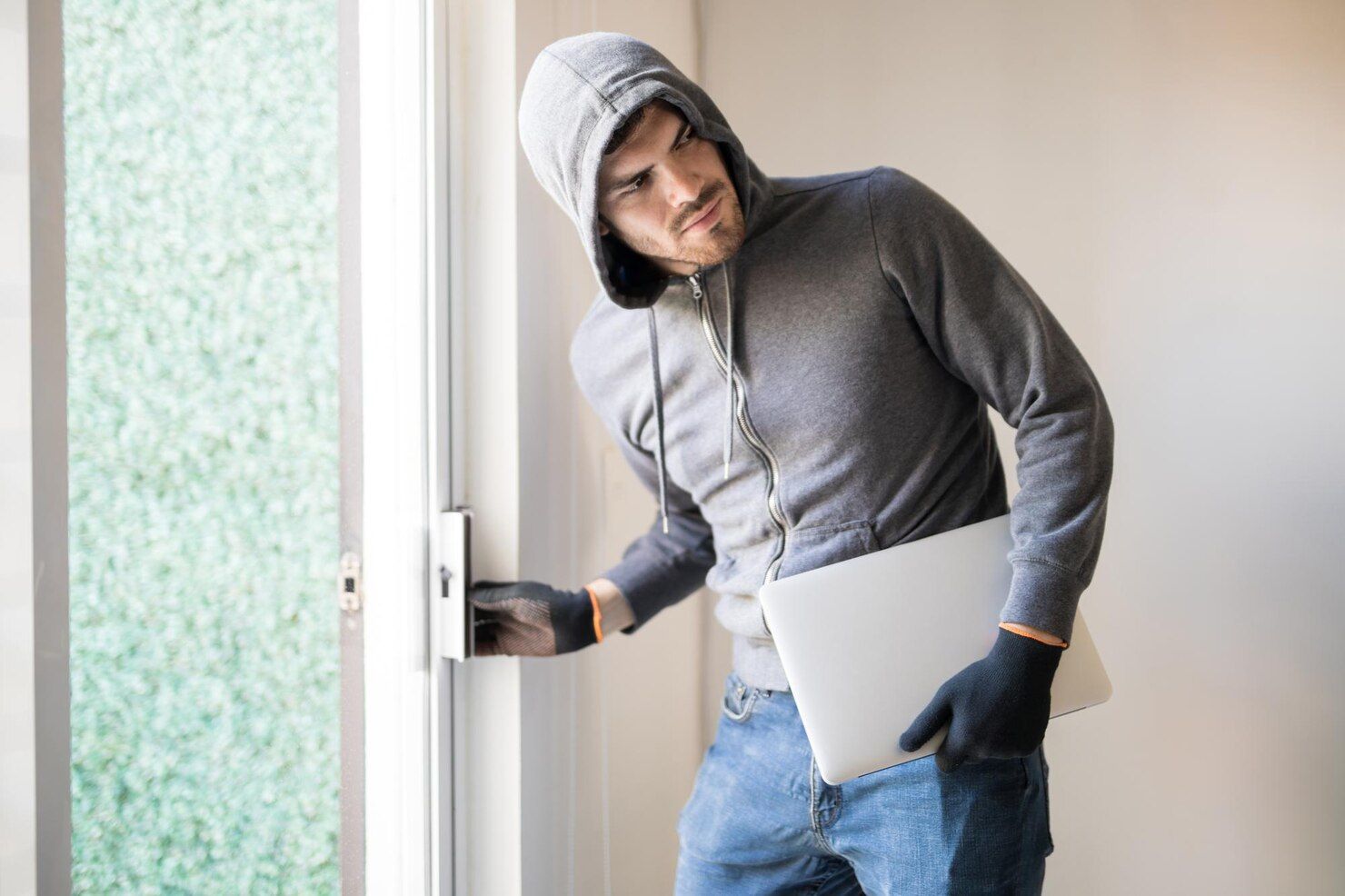Person in a hoody and gloves opens a door while holding a laptop; indoor setting. Burglary in Nashville, Tennessee