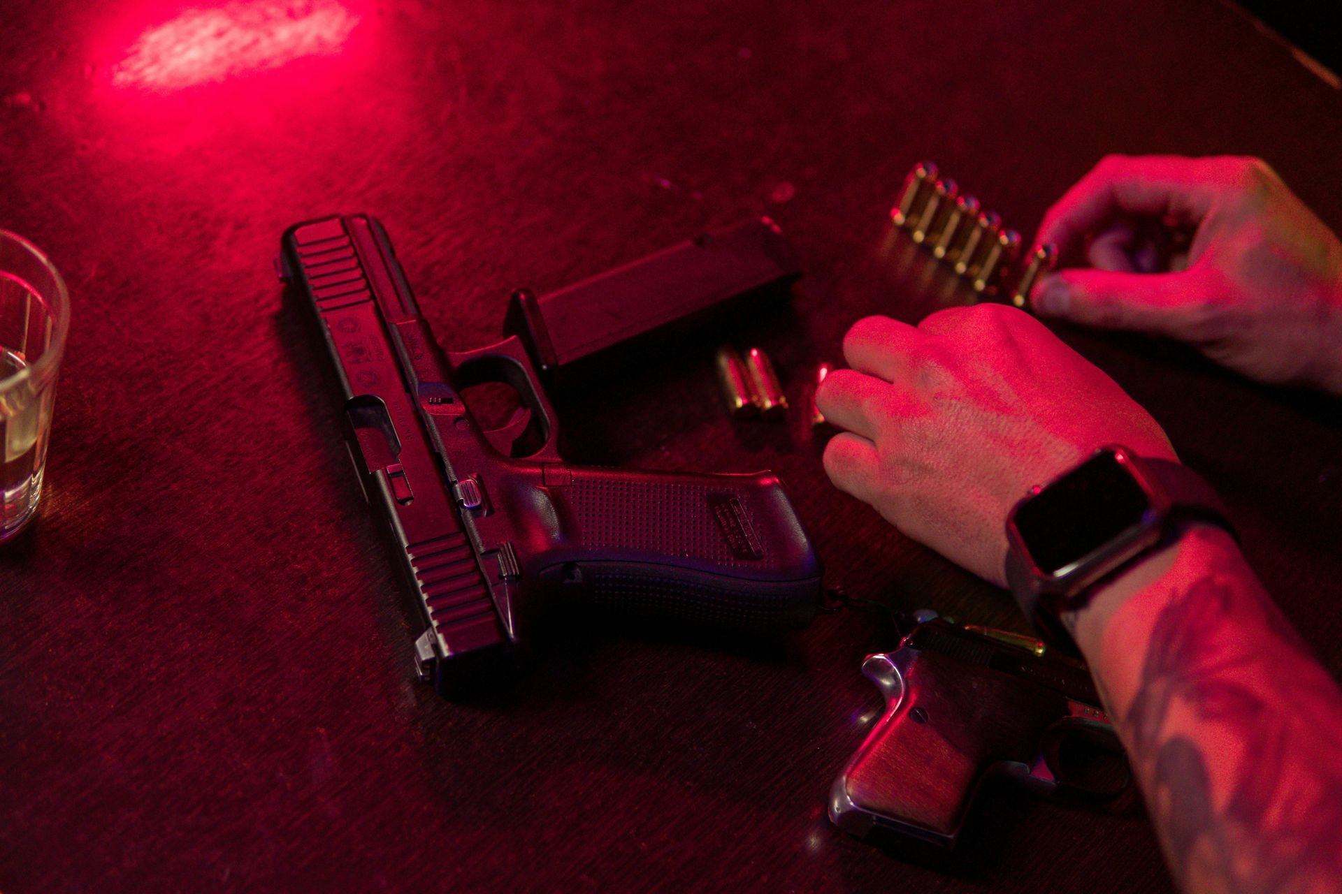A mans hands on a table where a handgun and ammo are on display in red lighting