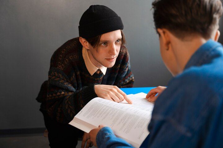 Person in beanie points to book held by another person. Dark sweater, blue shirt, gray wall. Second Degree Murder in Nashville, Tennessee