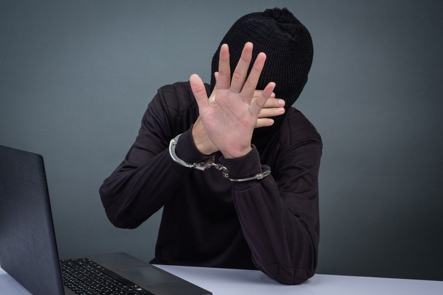 Person in a black balaclava and handcuffs, holding hands up near a laptop. Criminal Contempt in Nashville, Tennessee