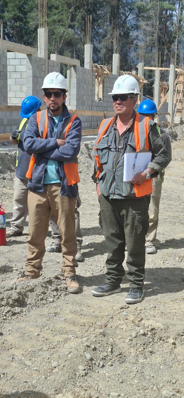 Un profesional con casco y chaleco reflectante sostiene planos en una obra de construcción con maquinaria cerca.
