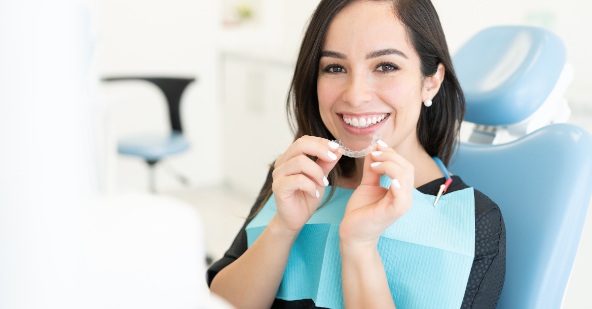Mulher sentada em cadeira de dentista, feliz, inserindo o alinhador invisível na boca