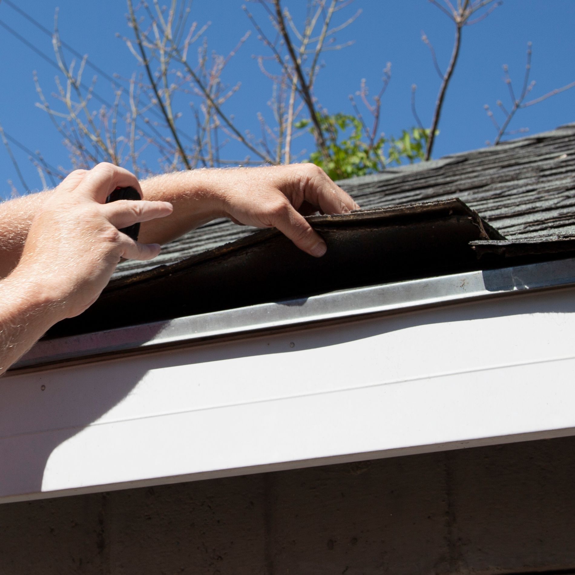 Hands repairing roof shingles near a gutter, outdoors under a blue sky.