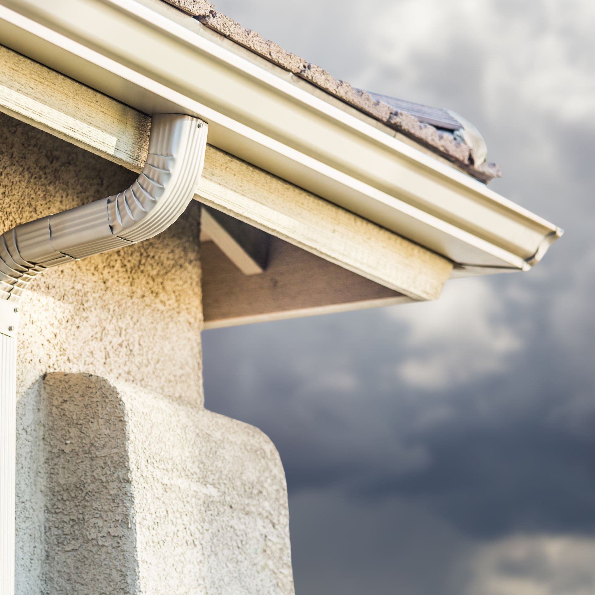 Beige gutter and downspout on a stucco house, against a cloudy sky.