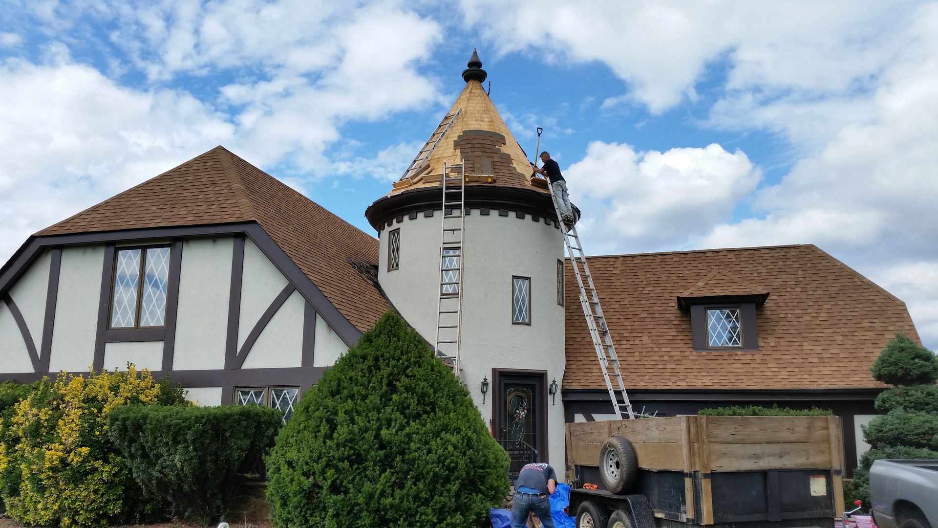 Workers on a roof of a Tudor-style home, with a tower. Blue sky, brown shingles, and trailer.