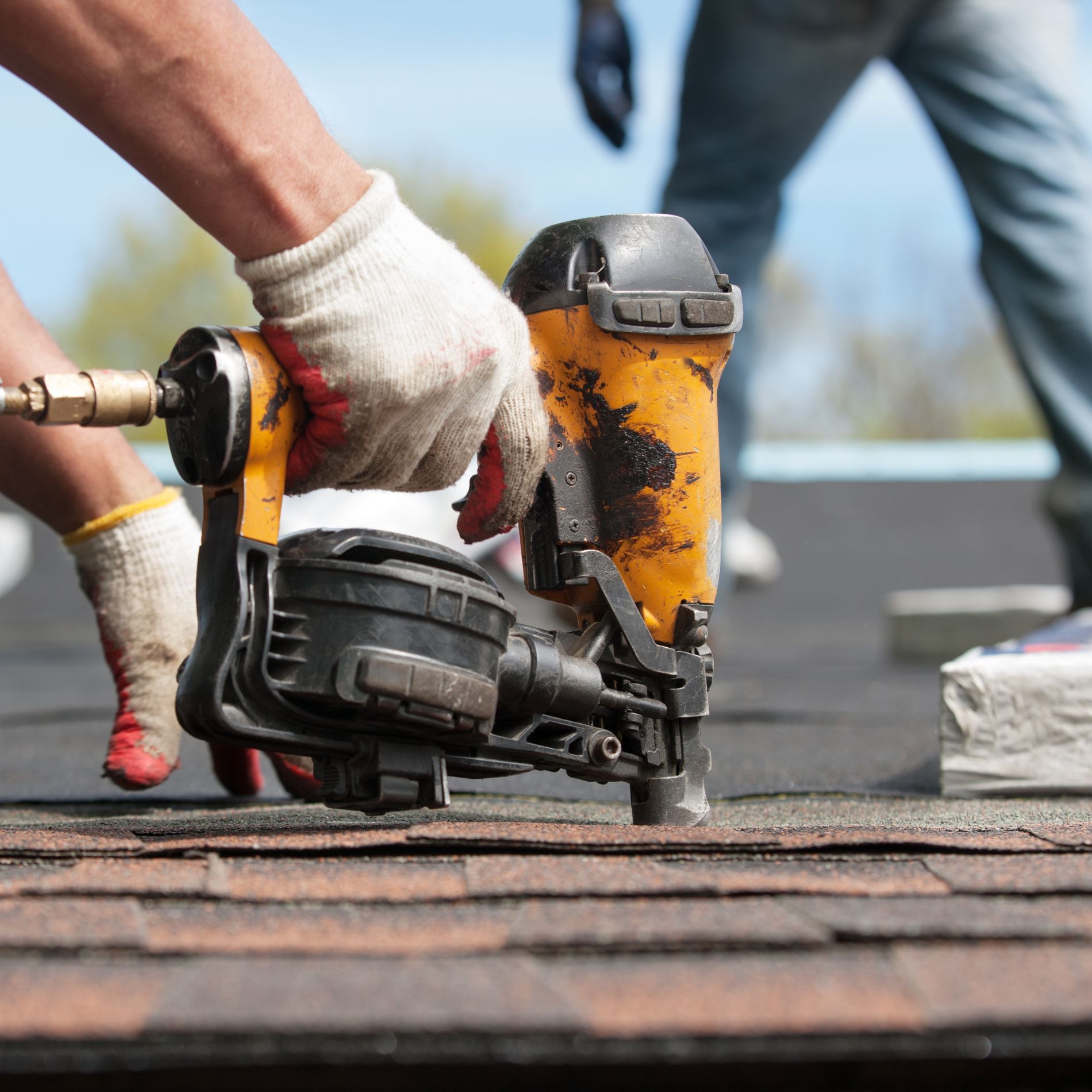 Roofer using a nail gun to install asphalt shingles on a roof, wearing gloves.