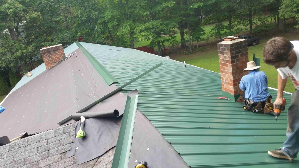 Workers installing a green metal roof. One is near a brick chimney, another is using a drill. 