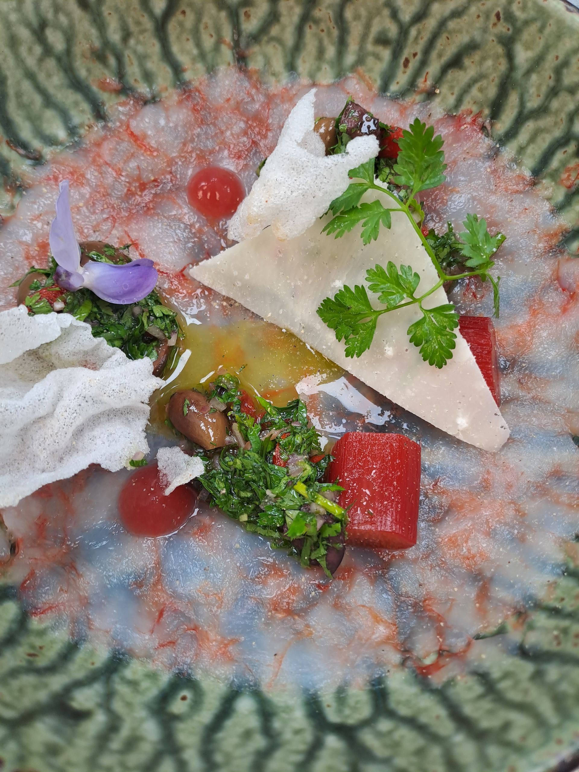 Deconstructed salad on green plate: white crisps, red and green chopped veggies, cheese triangle, and edible flower.