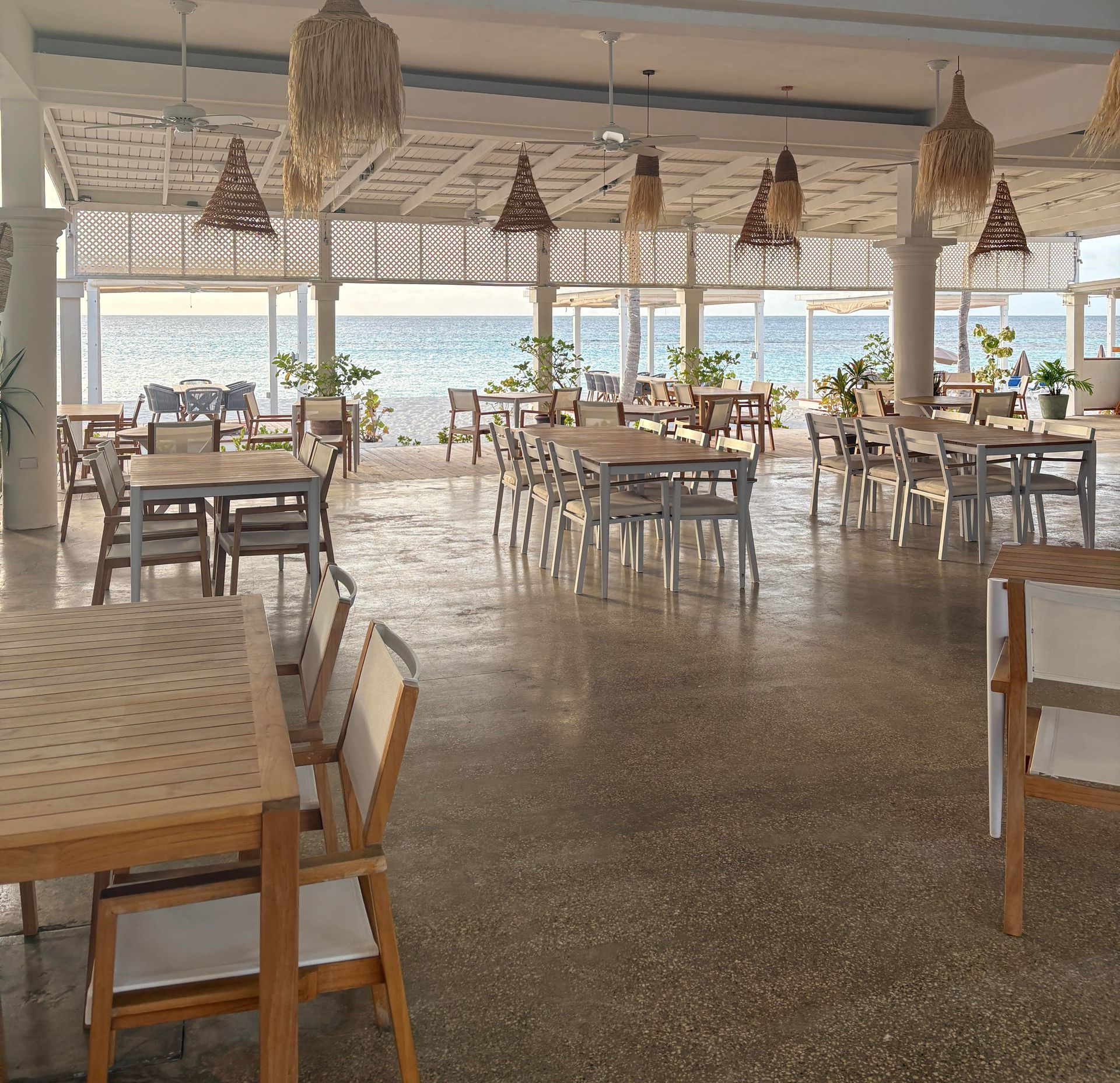 Empty restaurant with beach view, light wood tables, chairs, neutral tones, and hanging woven lamps.