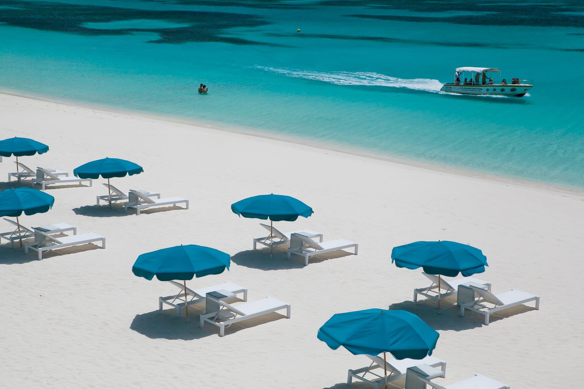 Beach scene with teal umbrellas, white lounge chairs, and turquoise water. A boat moves on the water.
