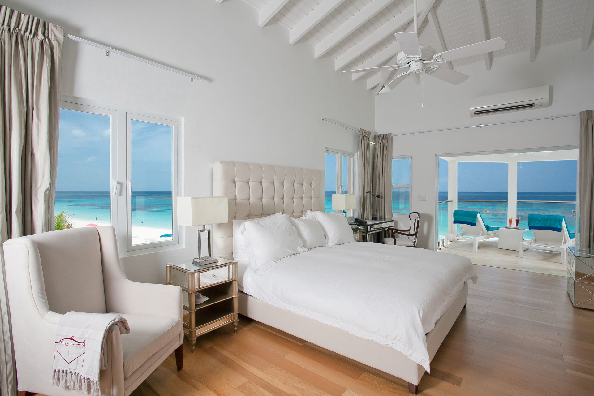 Bright, airy bedroom with beach view. White walls, wooden floor, large bed, armchair, and sliding doors to a deck.
