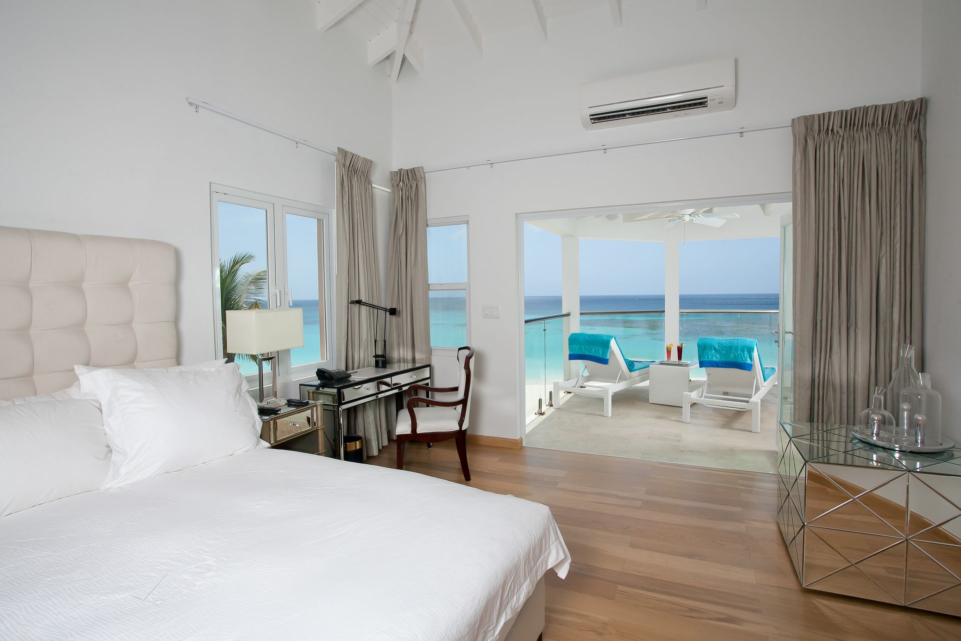 Bedroom with a bed, desk, and open doors leading to an ocean view and lounge chairs.
