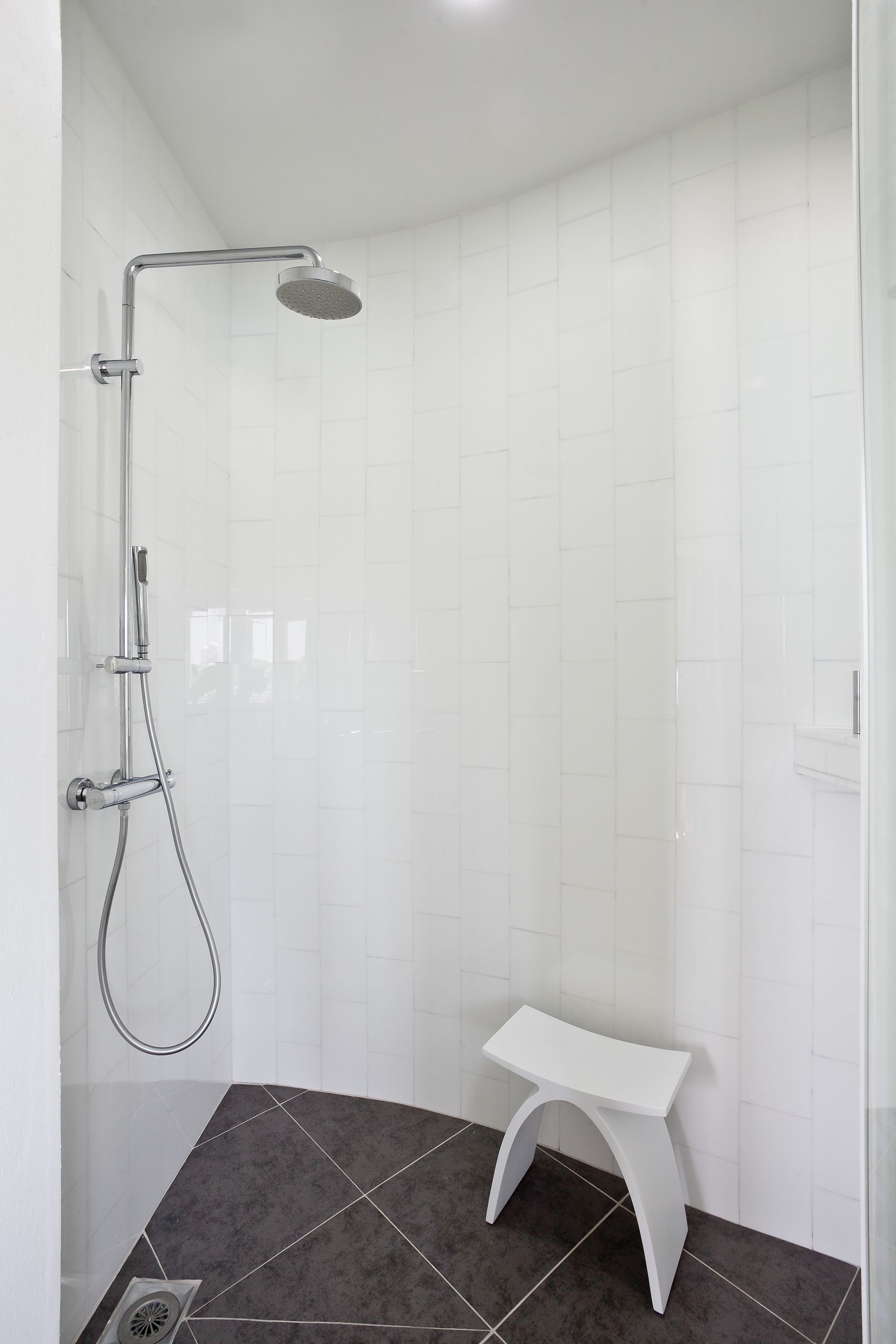 Shower with white tiled walls, dark tile floor, chrome fixtures, and a white stool.