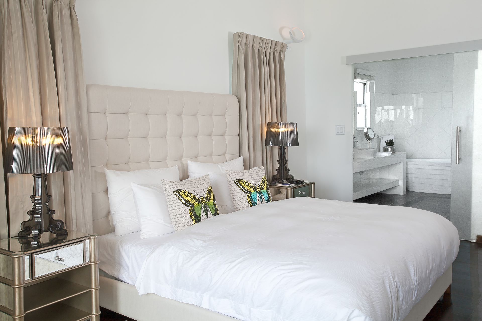 A bright bedroom with a white bed, mirrored nightstands, and sheer beige curtains. A glass door leads to the bathroom.