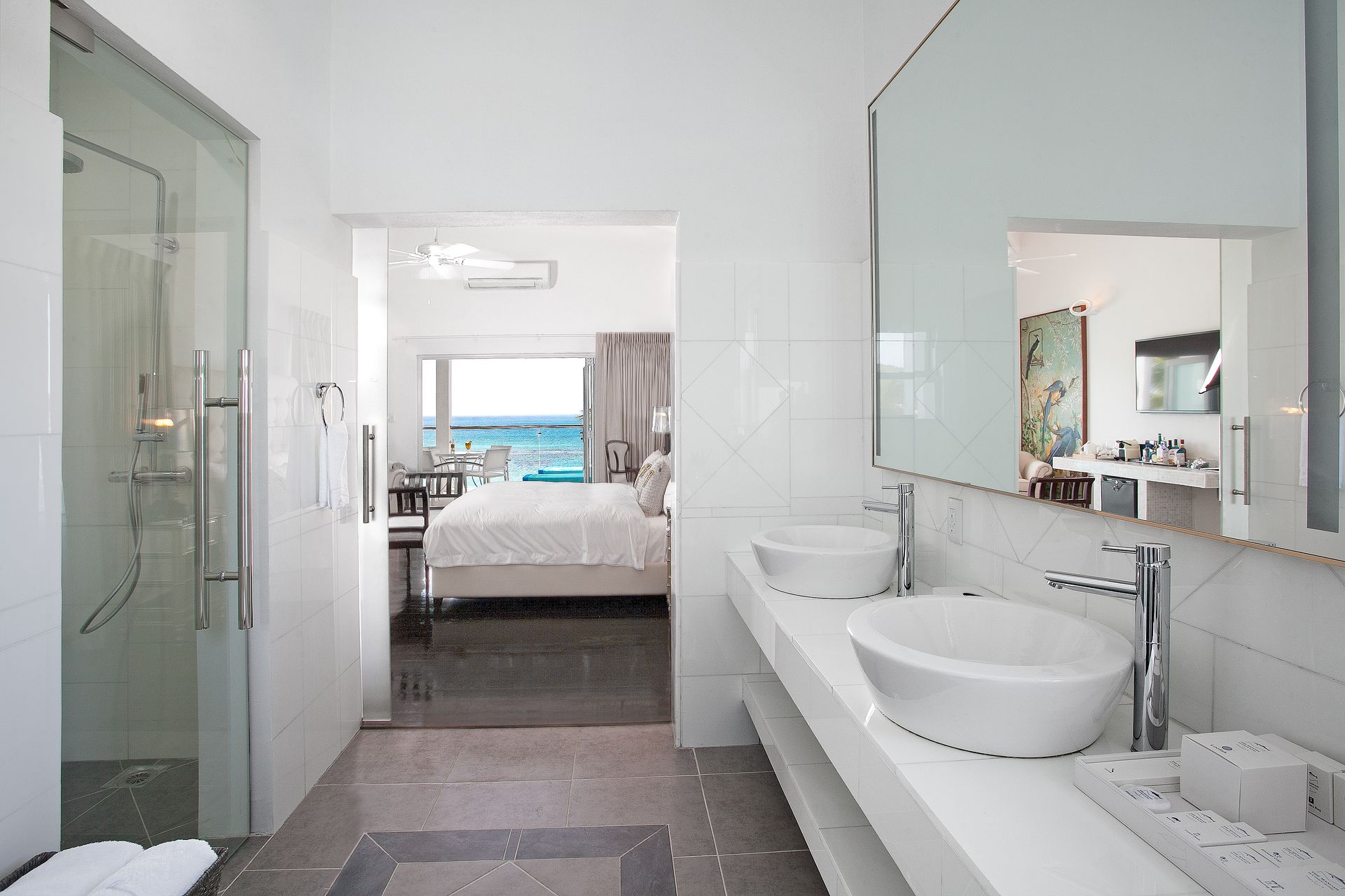 Modern white bathroom with shower, double sinks, and a view into a bedroom with a bed facing the ocean.