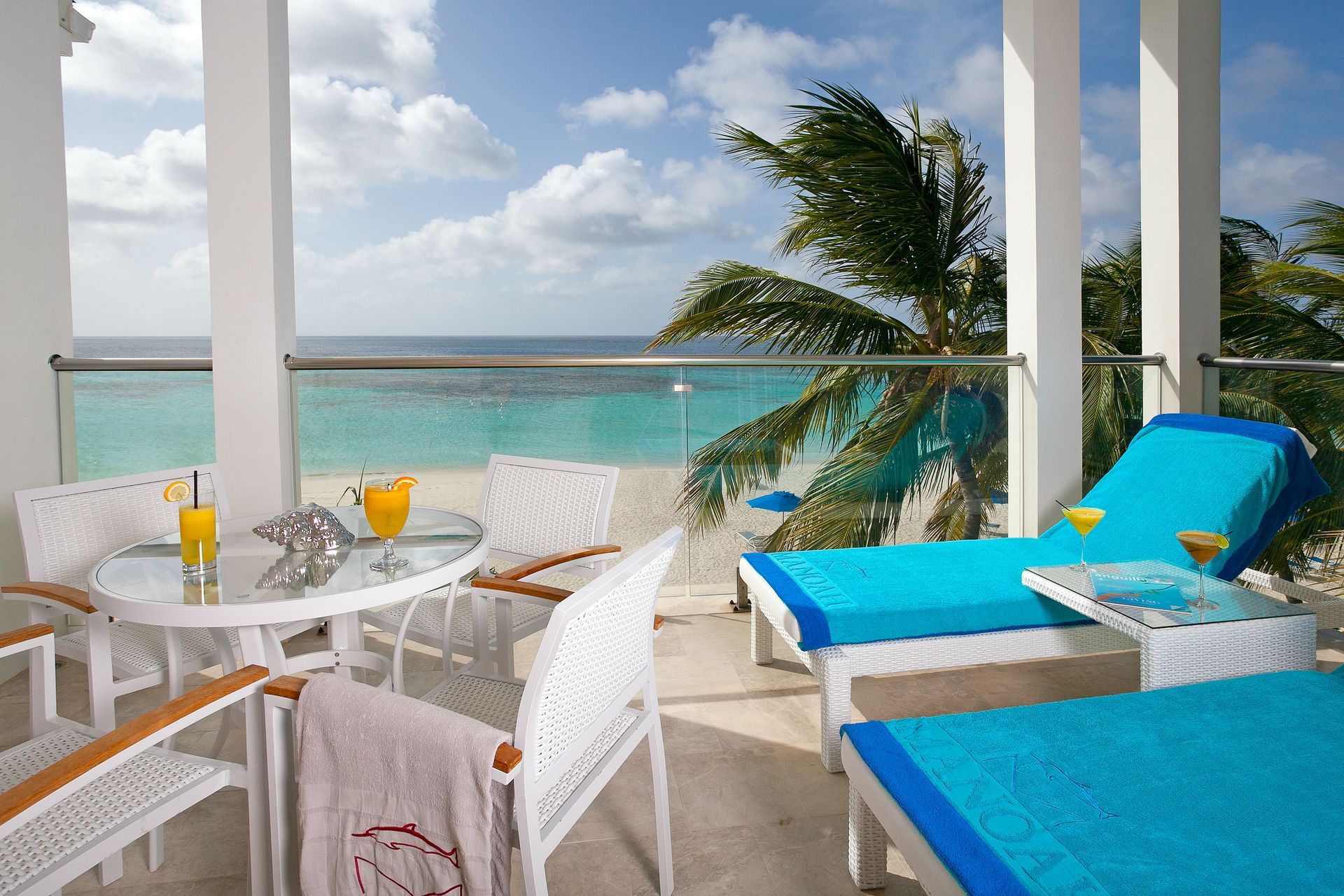 Balcony with ocean view: white furniture, turquoise lounge chairs, drinks, palm trees, blue sky.
