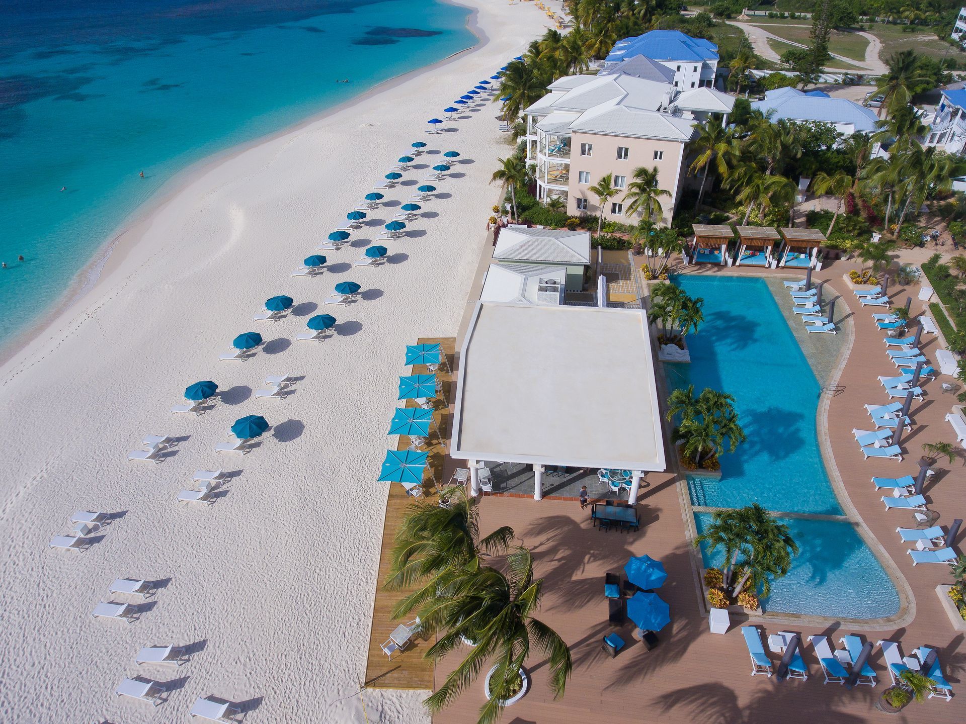 Aerial view of a tropical beach resort with white sand, turquoise water, and blue umbrellas.